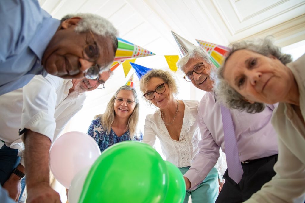friends-smiling-at-the-camera-7810931 Group of seniors celebrating indoors with balloons and party hats, sharing a joyful moment together.